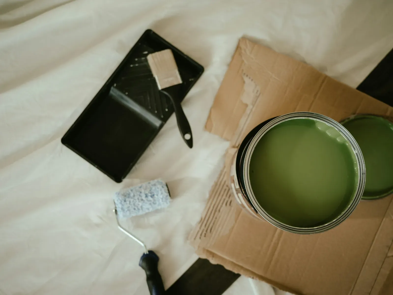An open tin of green paint with a roller and brush on a dust sheet